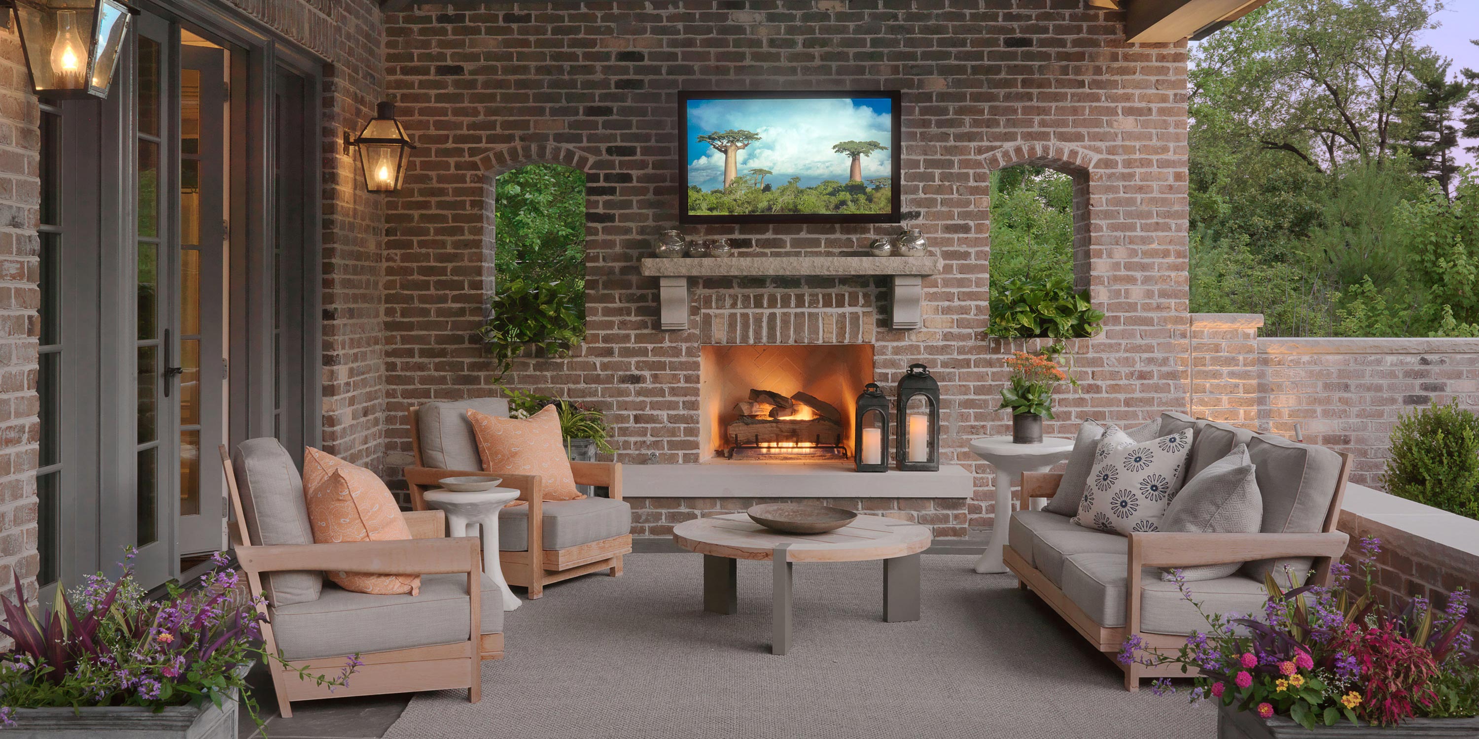 Cozy outdoor seating area with a brick fireplace, surrounded by greenery and warm lighting.