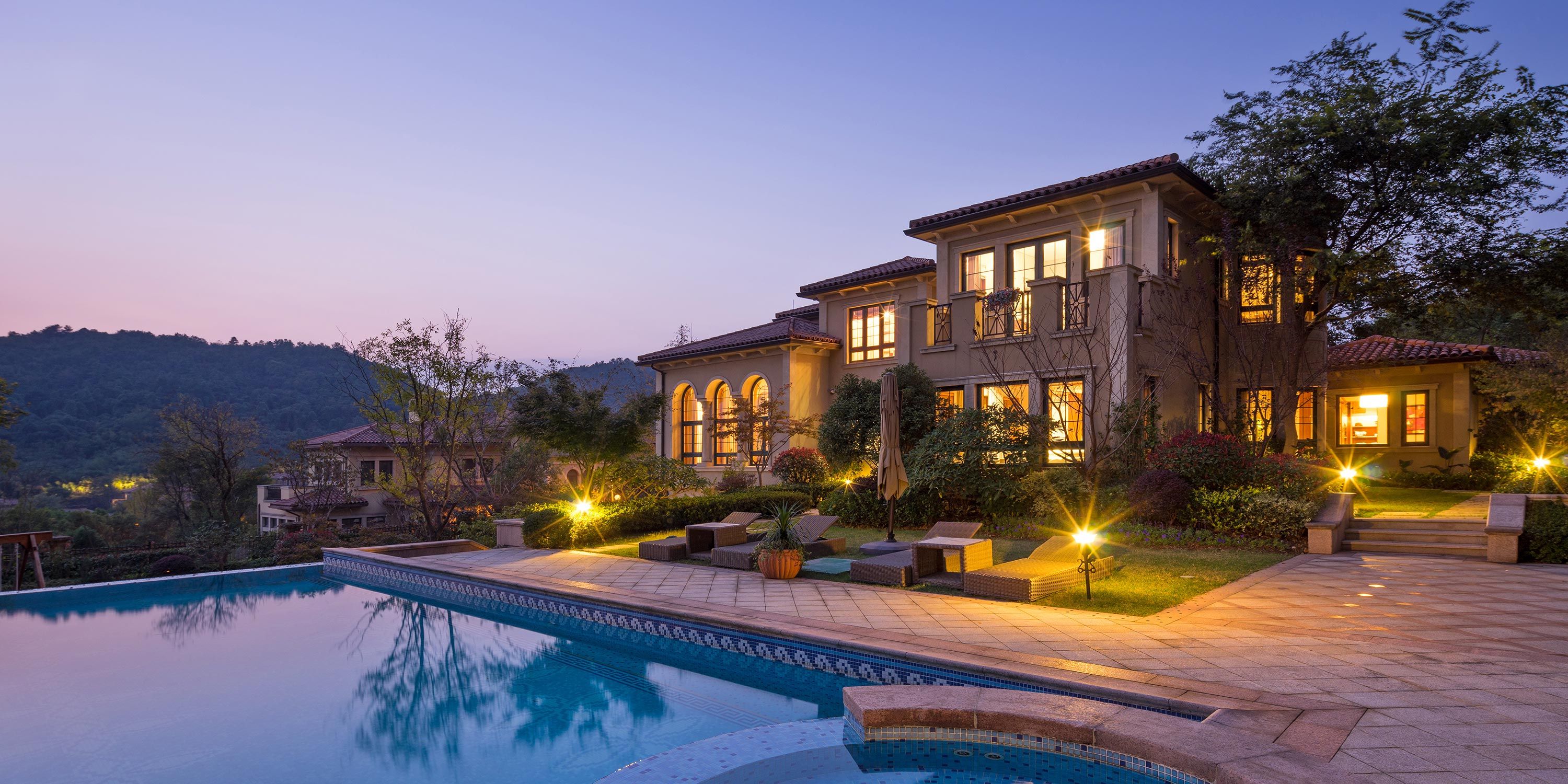 Elegant villa at twilight with a landscaped garden and poolside loungers, set against a hilly backdrop.