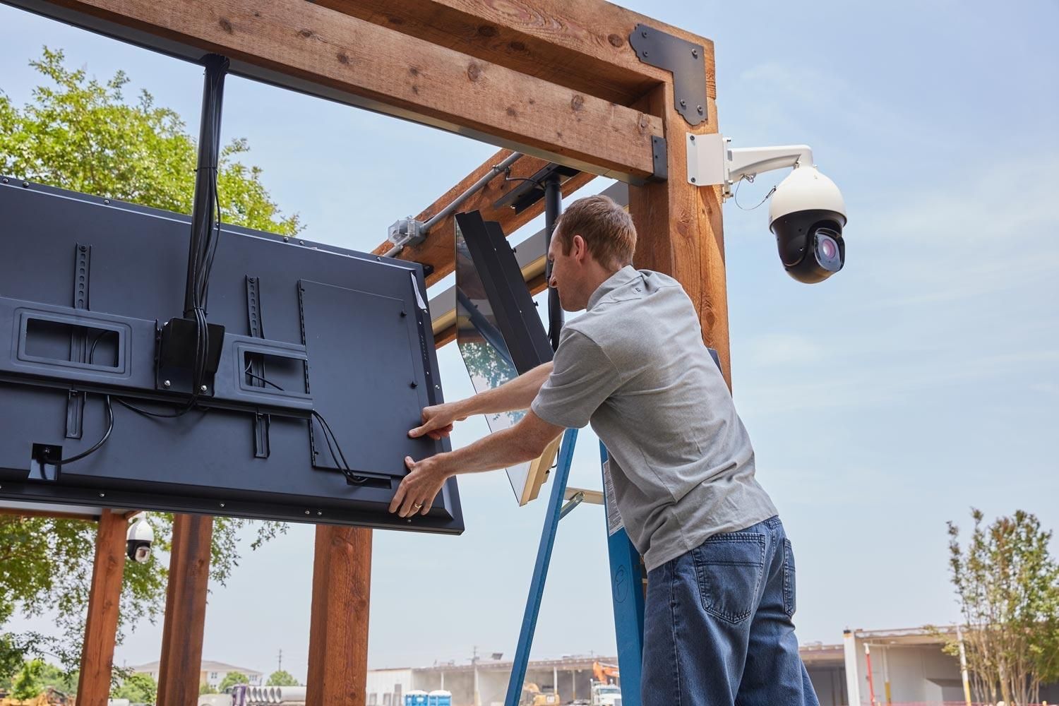 Man installing a large TV screen outdoors, with a security camera mounted on a wooden structure.