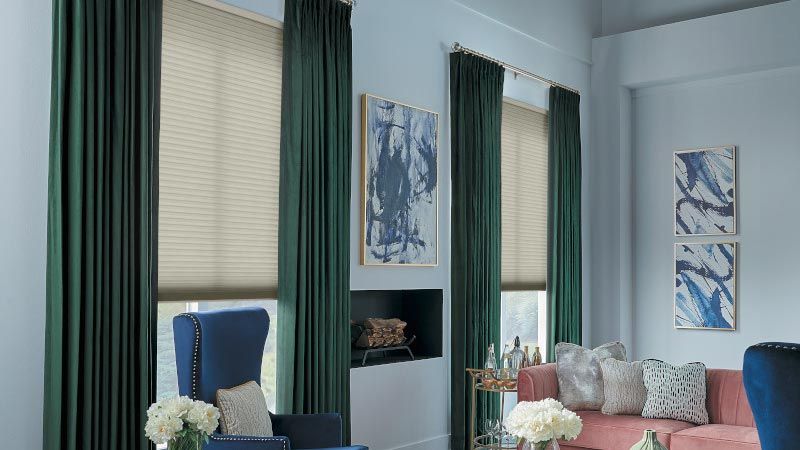 A stylish living room featuring dark green curtains, beige window shades, a blue wingback chair, and a pink sofa, with abstract art and floral arrangements adding a touch of elegance.