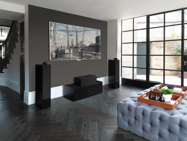 A modern living room featuring a wall-mounted TV, black speakers, a tufted gray ottoman, and large windows with black frames.