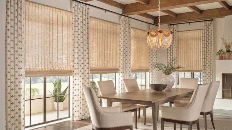 A cozy dining room with a wooden table and beige chairs, surrounded by windows with woven shades and patterned curtains, and adorned with a decorative chandelier and greenery.