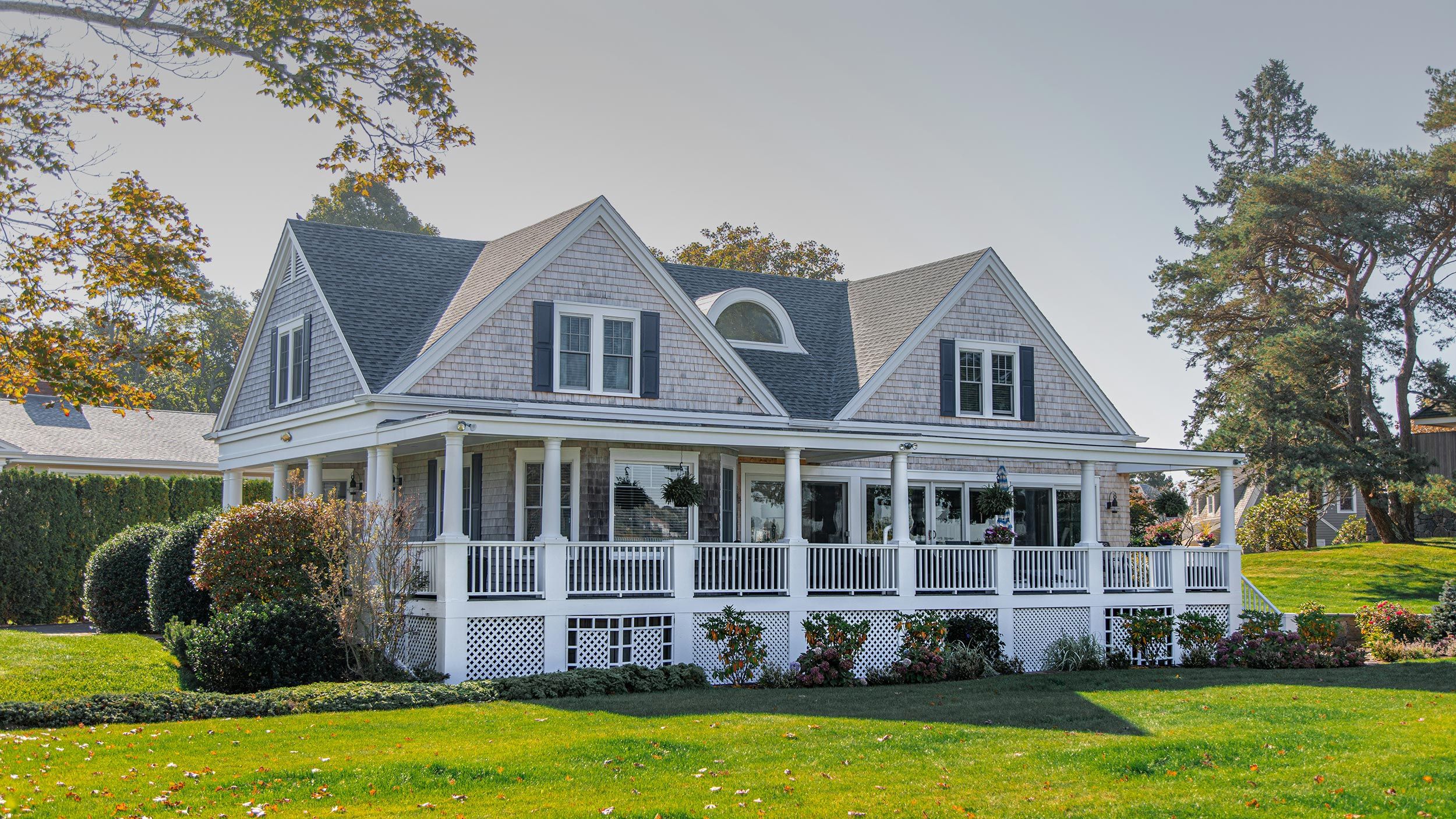 Beautiful two-story house with a wraparound porch, grey shingles, and a well-manicured lawn.