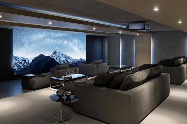 A modern home theater with gray seating and a large screen displaying a mountain scene, illuminated by recessed lighting.
