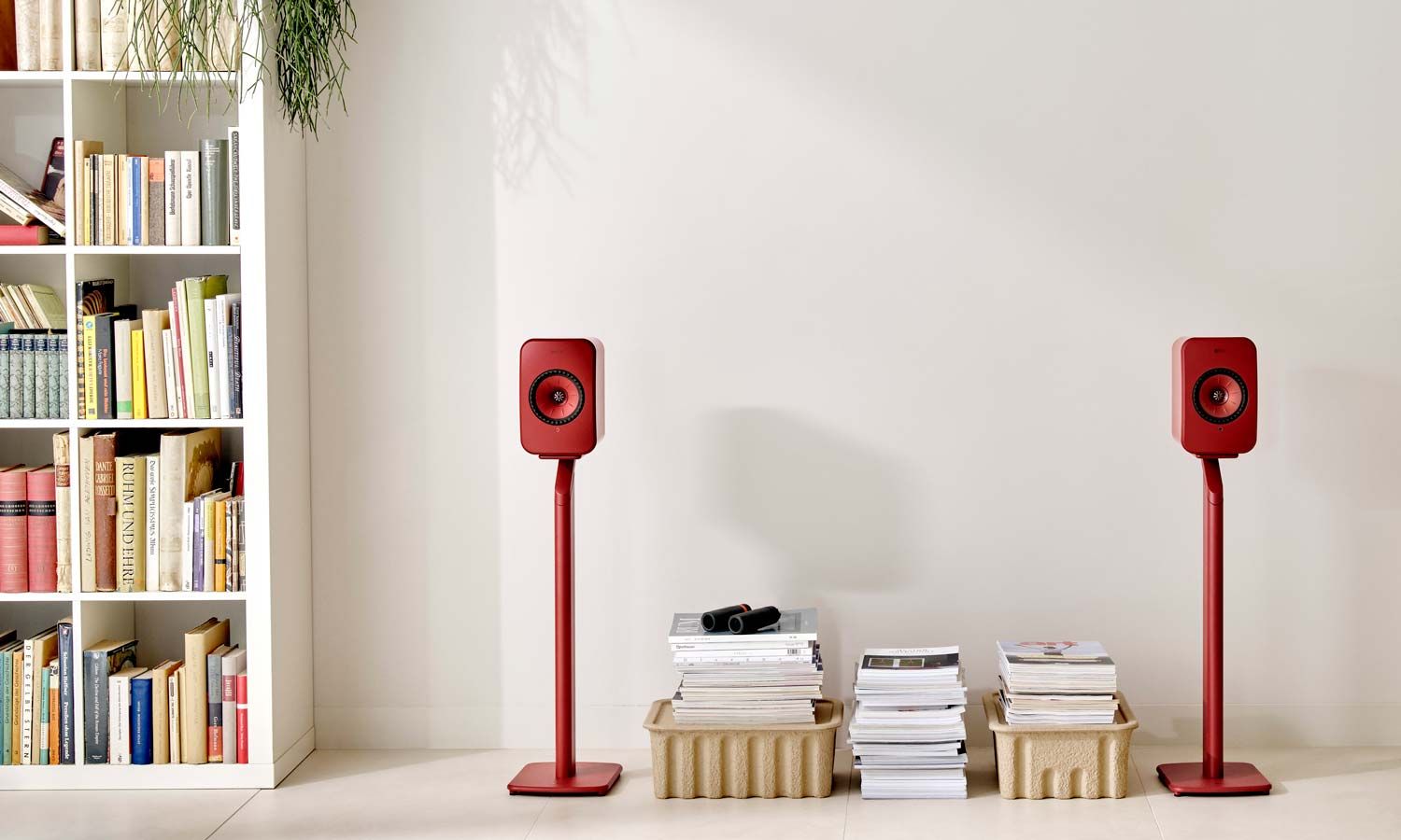 A pair of red KEF LSX II speakers on stands in a cozy room with a bookshelf.