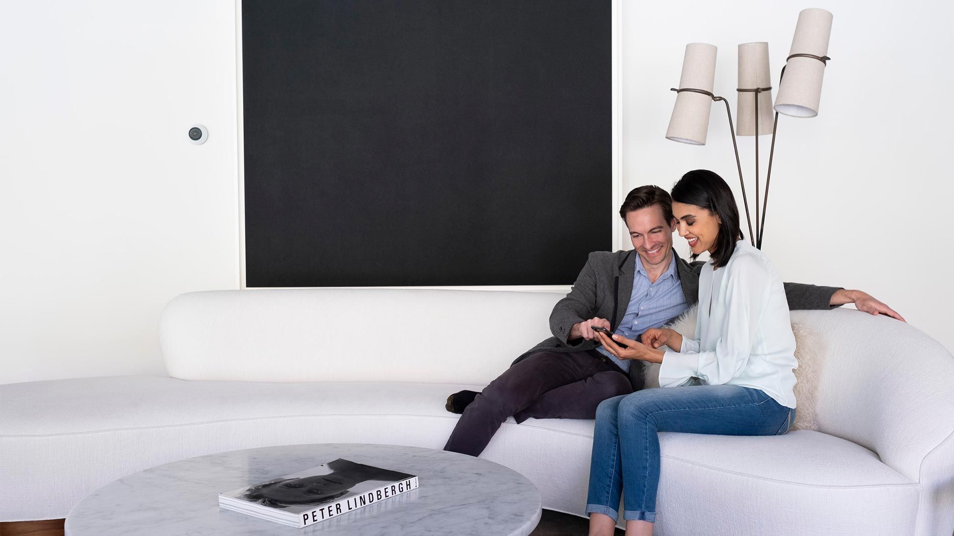 Couple sitting on a white couch, smiling while looking at a tablet in a modern living room.