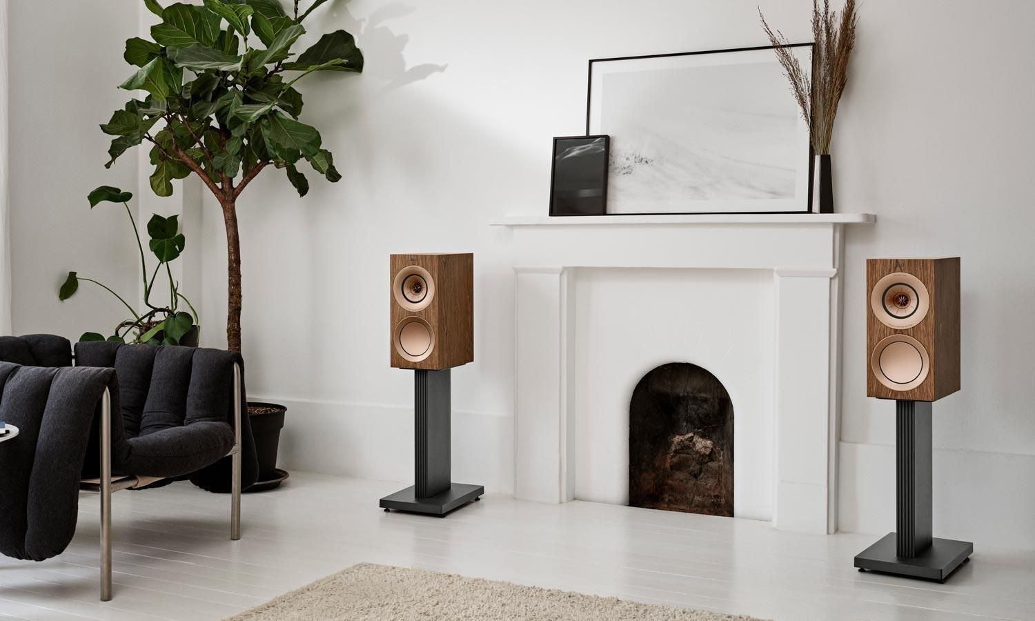 A pair of KEF R Series bookshelf speakers in a wooden finish on stands in a bright, modern living room.