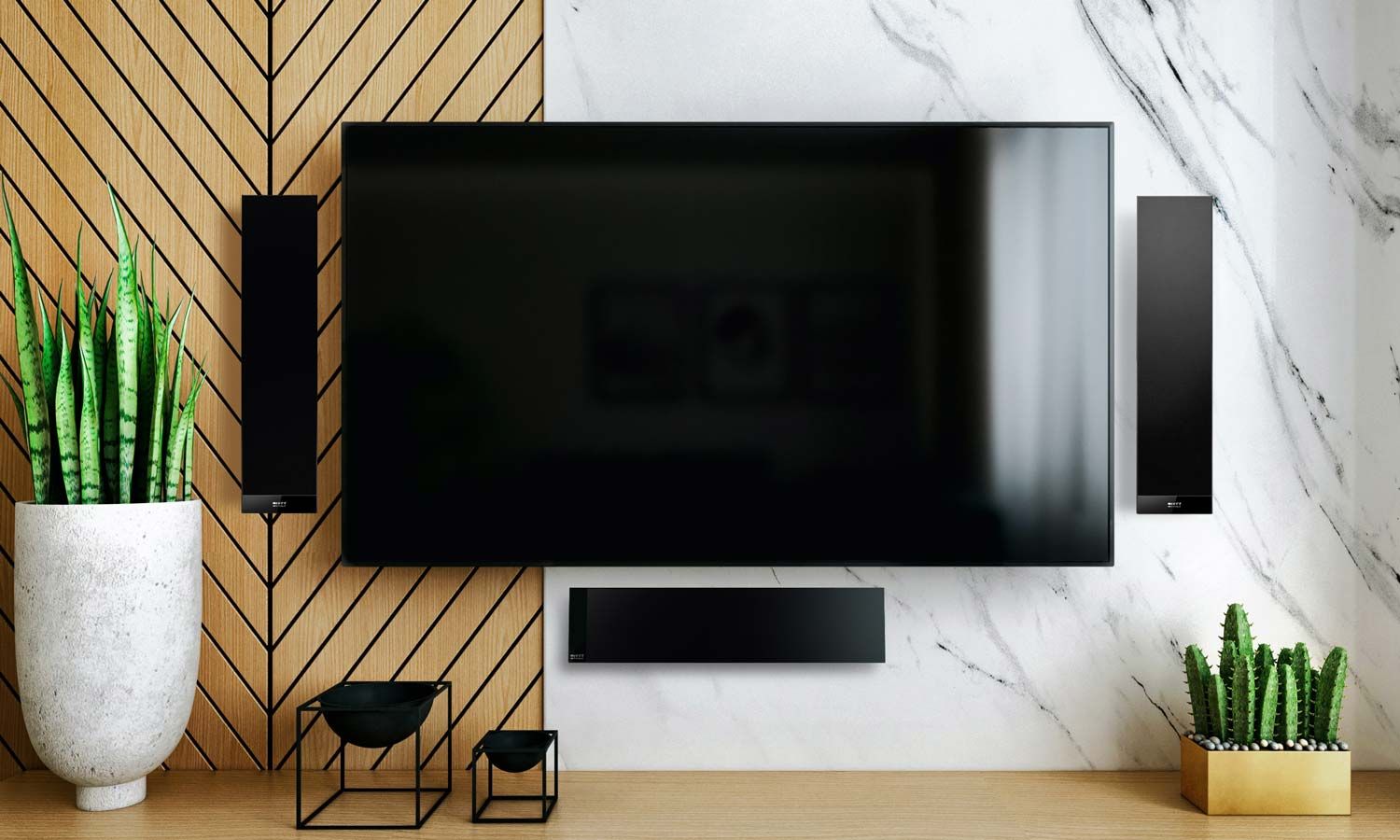 A stylish modern entertainment setup featuring a flat-screen TV flanked by two vertical KEF Audio speakers on a wood and marble accent wall, with decorative plants and minimalist black metal bowls on a wooden console.