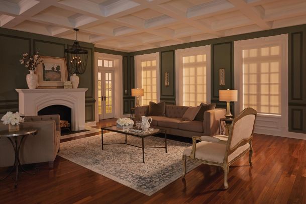 A cozy living room with dark green walls, white trim, and a coffered ceiling, featuring a brown couch, armchair, and a white fireplace.