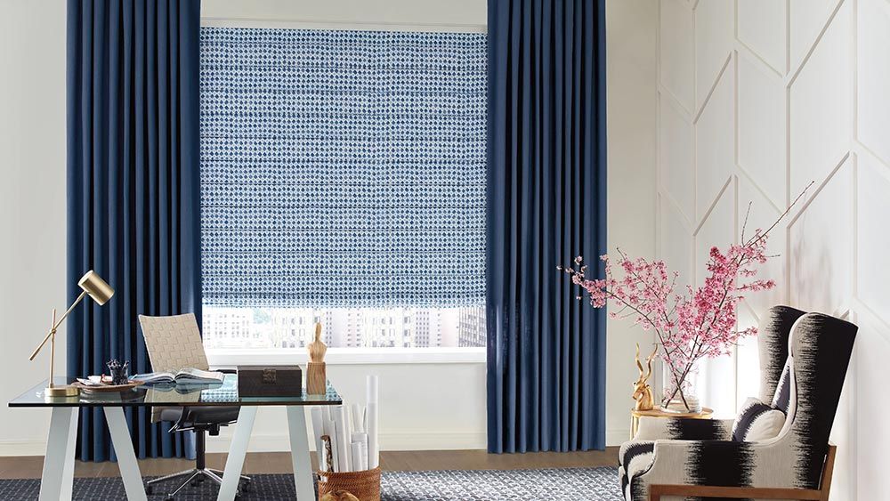 A sophisticated office space with a glass desk, blue patterned window shades, and dark blue curtains, complemented by a modern black chair and a decorative branch with pink flowers.