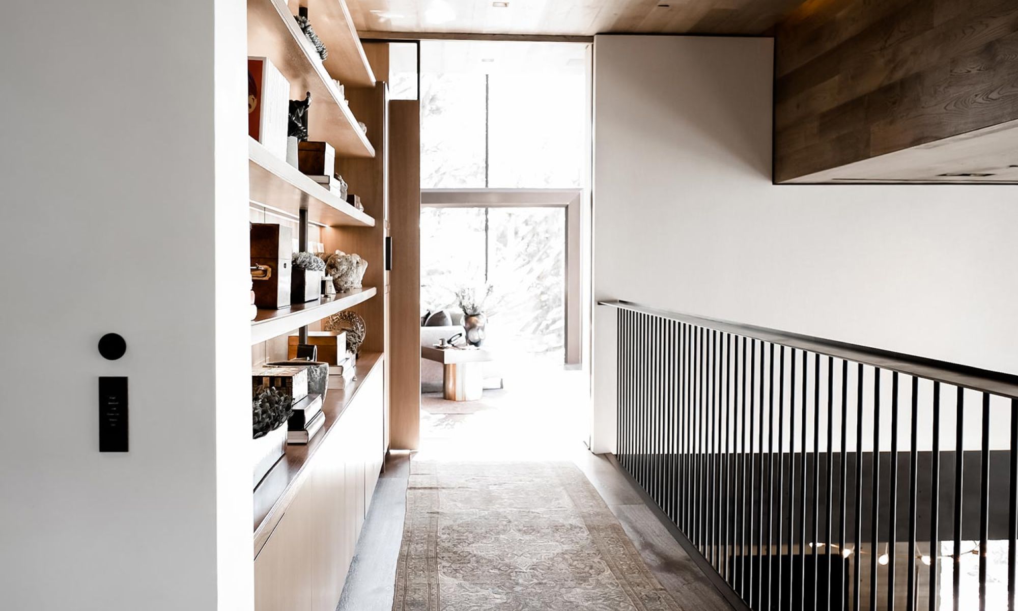 A modern hallway featuring built-in wooden shelves with decorative items and a railing with vertical black bars, leading to a bright living area with large windows. On the wall a Josh Nano device in black.