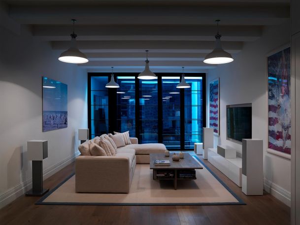A sleek living room with a white sectional sofa, white square speakers, and large windows with a view of the city, lit by pendant lights.