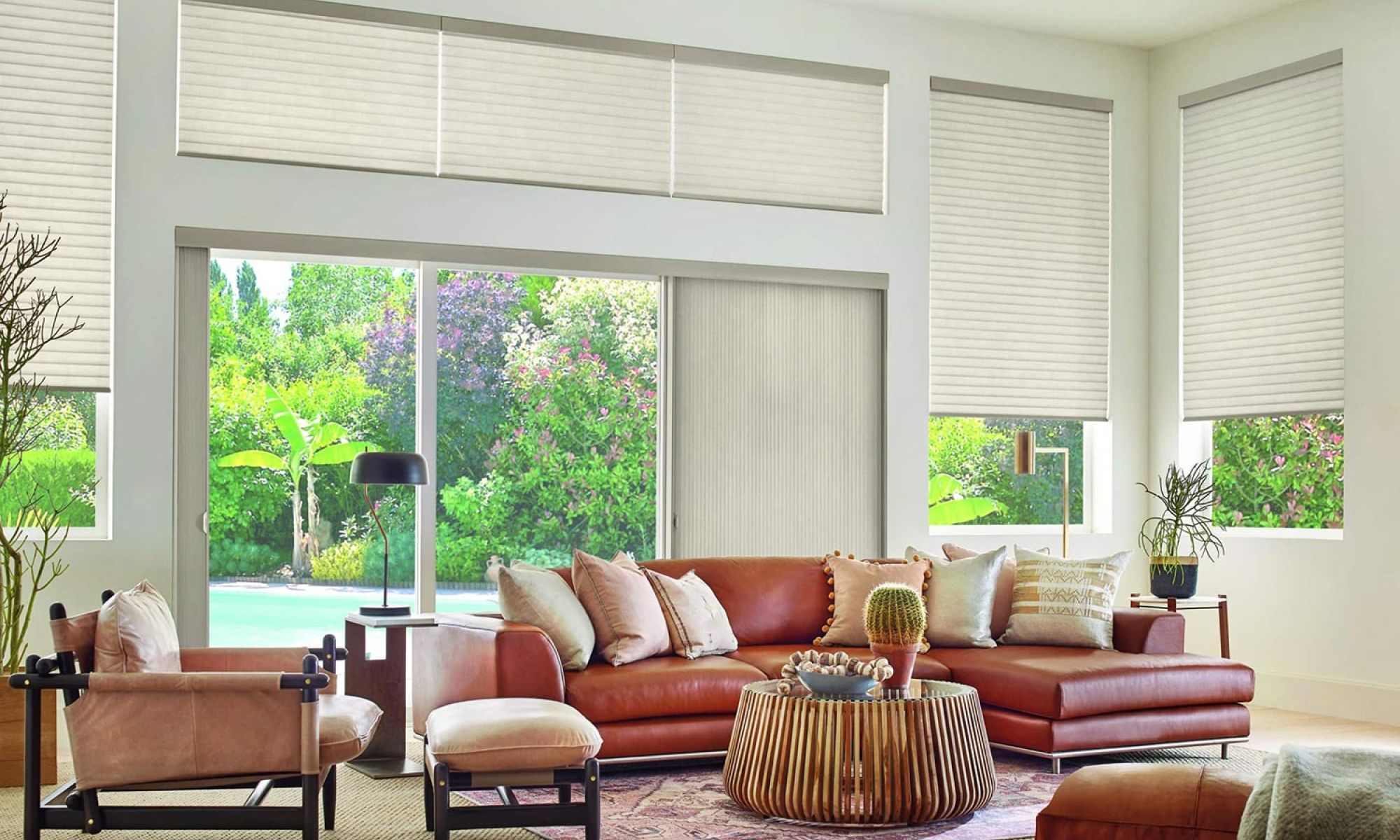 A bright and cozy living room with large windows with Hunter Douglas shades showcasing a lush garden view, featuring a brown leather sectional sofa, matching armchairs, and a round wooden coffee table adorned with a cactus and decorative items.