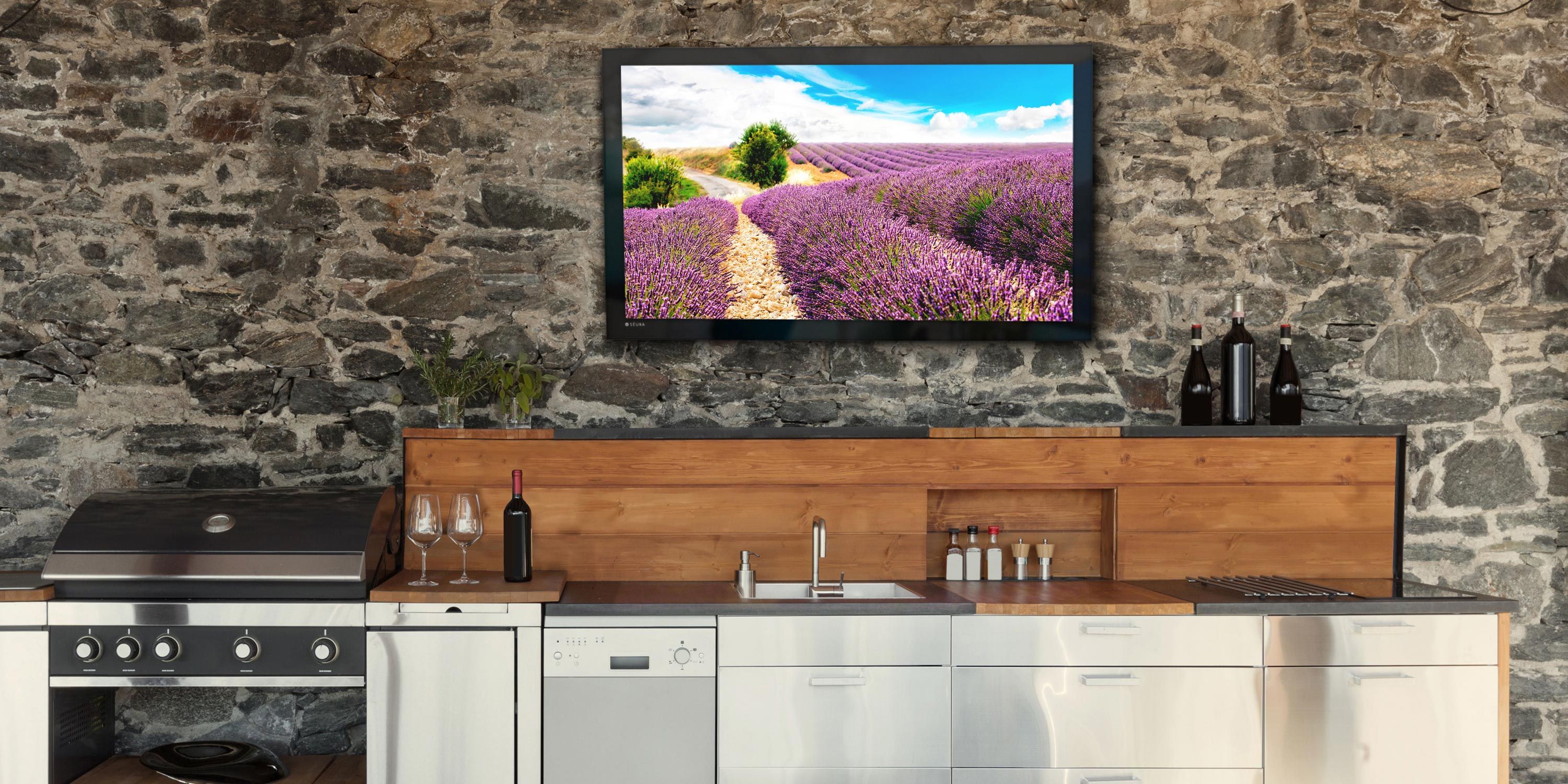 Rustic outdoor kitchen with a stone wall, stainless steel appliances, and a vibrant lavender field on the TV.