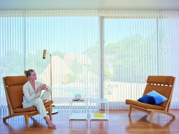 A bright living room with floor-to-ceiling windows covered by sheer curtains, two orange armchairs, and a small white table.