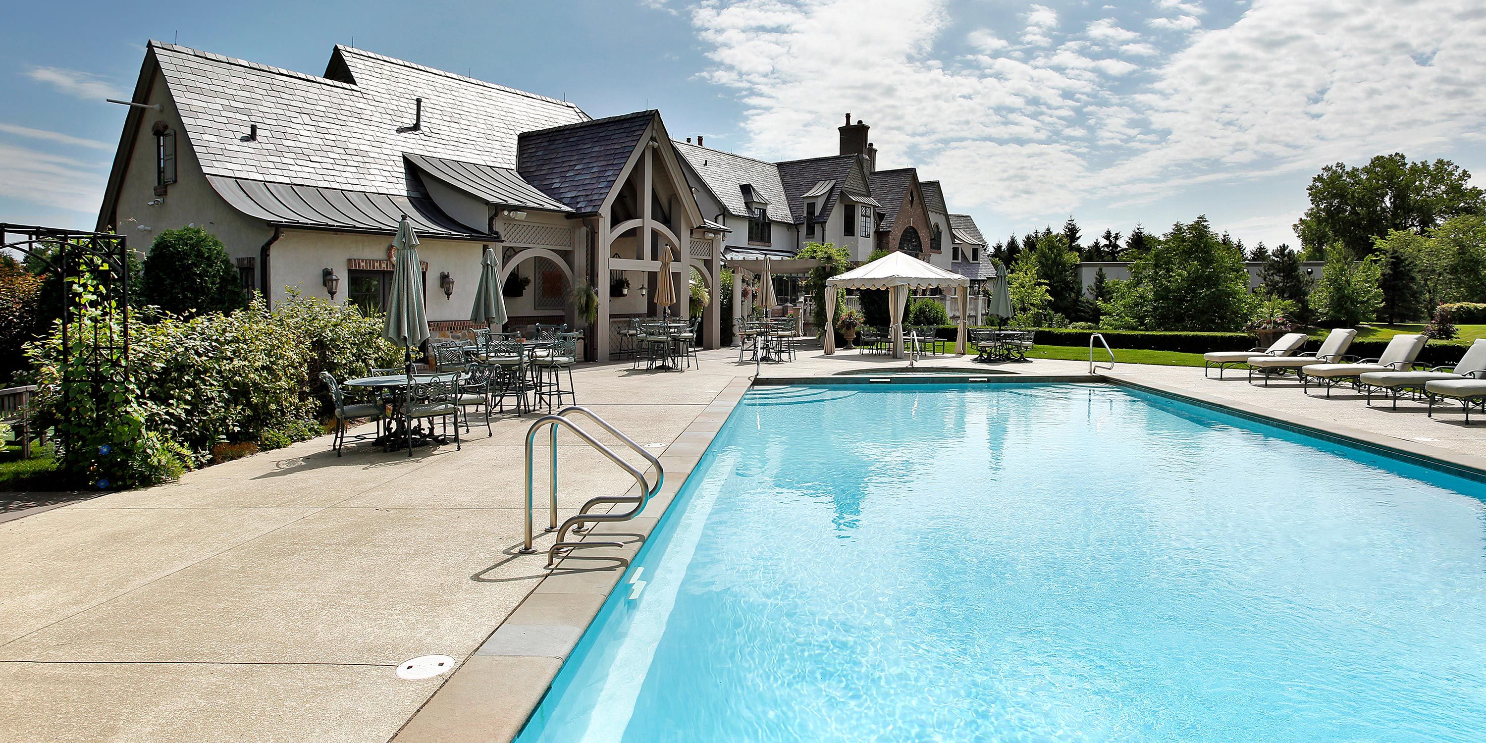 Spacious pool area with a luxurious house, surrounded by greenery and poolside seating.