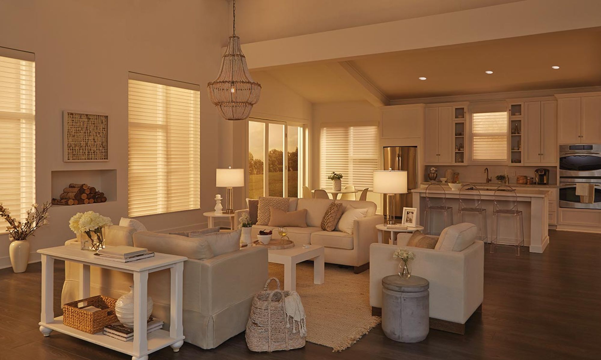 A warm, inviting open-concept living and dining area with cream-colored furniture, large windows with Lutron blinds, and a chandelier, creating a cozy atmosphere during sunset.