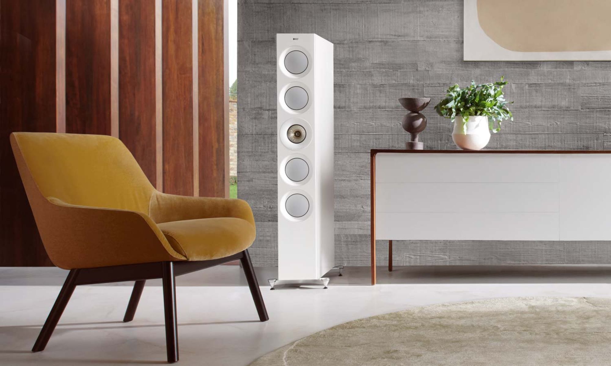 A contemporary living room setup with a mustard yellow chair, a white KEF Audio speaker tower, and a white sideboard with decorative plants against a textured gray wall.