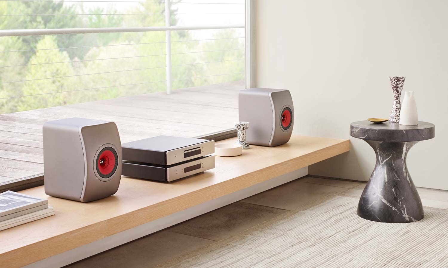 A pair of grey KEF LS50 Meta speakers with red drivers displayed on a light wooden shelf in a modern living room setting.